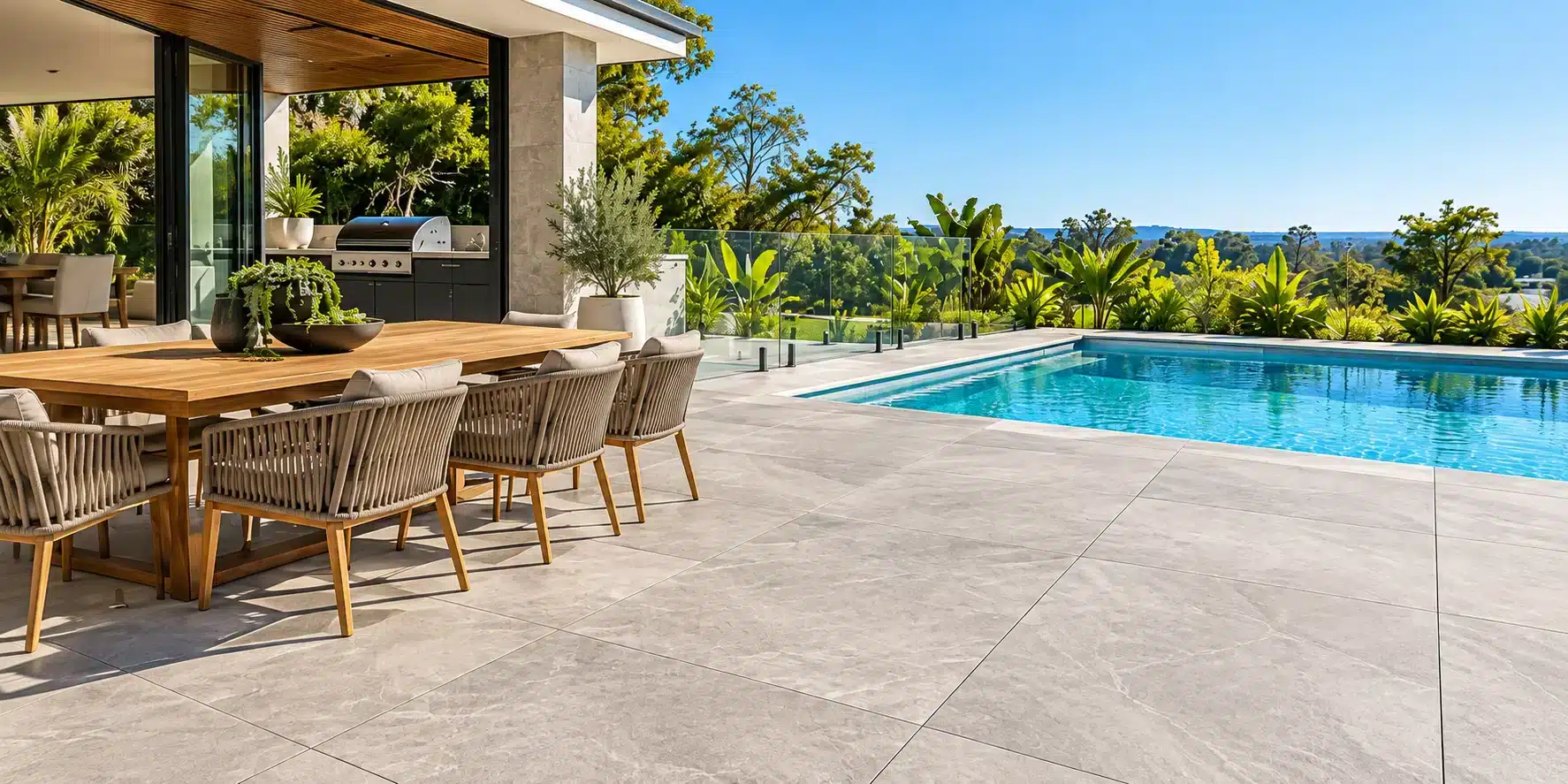 Grey porcelain paving across a sunny poolside patio with an outdoor dining table and kitchen.